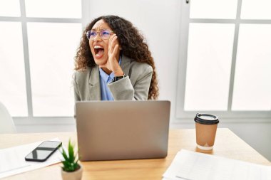 Güzel bir İspanyol iş kadını ofiste oturmuş dizüstü bilgisayarla çalışıyor ve elleri ağızda bağırıyor. iletişim konsepti. 