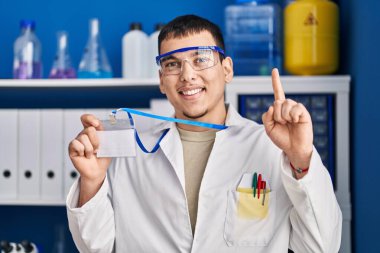 Young arab man working at scientist laboratory holding id card smiling with an idea or question pointing finger with happy face, number one 