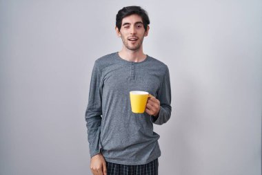 Young hispanic man wearing pajama drinking a cup of coffee winking looking at the camera with sexy expression, cheerful and happy face. 
