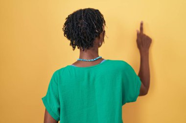 African woman with dreadlocks standing over yellow background posing backwards pointing ahead with finger hand 