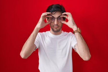Young hispanic man standing over red background trying to open eyes with fingers, sleepy and tired for morning fatigue 