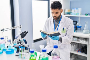 Laboratuvarda bilim adamı üniforması giyen genç İspanyol adam kitap okuyor.