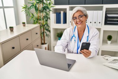 Klinikte doktor üniforması giyen orta yaşlı bir kadın akıllı telefon kullanıyor.