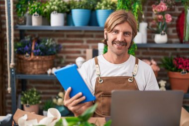 Çiçekçide dizüstü bilgisayar ve dokunmatik ped kullanarak gülümseyen genç sarışın adam.