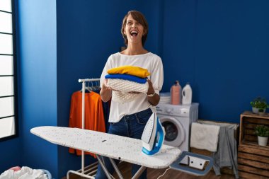 Brunette woman holding folded laundry after ironing angry and mad screaming frustrated and furious, shouting with anger looking up. 