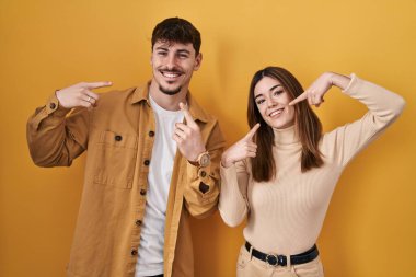 Young hispanic couple standing over yellow background smiling cheerful showing and pointing with fingers teeth and mouth. dental health concept. 