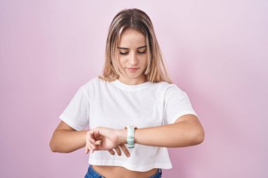 Young blonde woman standing over pink background checking the time on wrist watch, relaxed and confident 