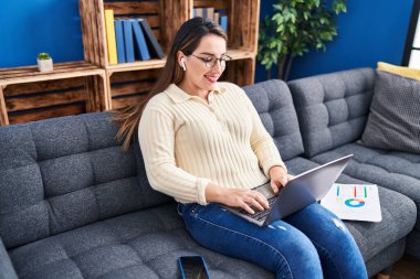 Young beautiful hispanic woman using laptop and wireless earphones sitting on sofa at home