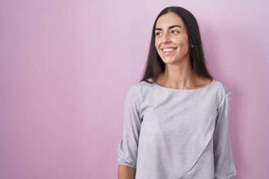 Pembe arka planda duran genç esmer kadın yüzünde gülümsemeyle yan yana bakıyor, doğal bir ifade. Kendinden emin gülen. 