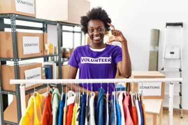 African young woman wearing volunteer t shirt at donations stand smiling and confident gesturing with hand doing small size sign with fingers looking and the camera. measure concept. 