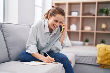 Not defterine yazan genç bir kadın evde akıllı telefondan konuşuyor.