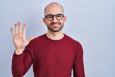 Beyaz arka plan üzerinde duran sakallı genç bir adam gözlük takıyor ve beş numaralı parmaklarıyla işaret ederken kendinden emin ve mutlu gülümsüyor.. 