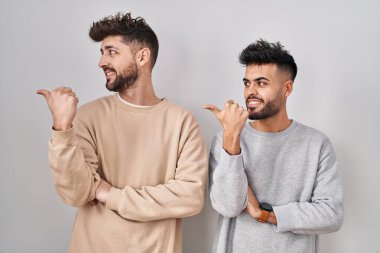 Young homosexual couple standing over white background smiling with happy face looking and pointing to the side with thumb up. 