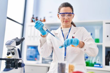 Young beautiful hispanic woman scientist pouring liquid on sample at laboratory