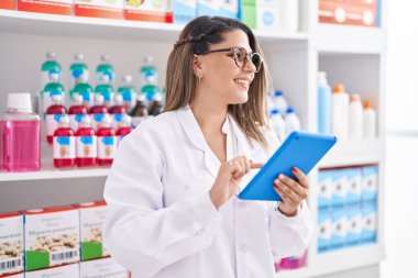 Young hispanic woman pharmacist smiling confident using touchpad at pharmacy
