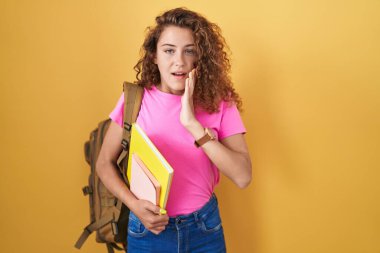 Öğrenci çantası takan, elinde kitap tutan, korkmuş, şok olmuş, şaşırmış ve şaşırmış genç beyaz bir kadın. 