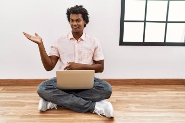African man with curly hair using laptop sitting on the floor smiling cheerful presenting and pointing with palm of hand looking at the camera. 