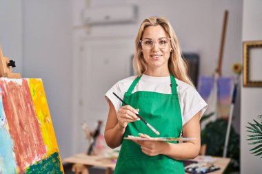 Sanat stüdyosunda kendine güvenen genç sarışın sanatçı gülümsüyor.