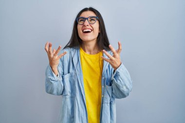 Mavi arka planda dikilen Latin kökenli genç kadın kızgın ve deli gibi bağırıyor ve agresif bir ifade ve kollarla bağırıyor. hüsran kavramı. 