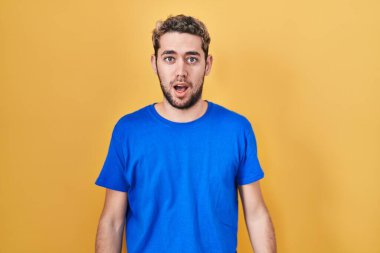 Hispanic man with beard standing over yellow background afraid and shocked with surprise and amazed expression, fear and excited face. 