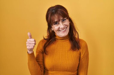 Middle age hispanic woman standing over yellow background doing happy thumbs up gesture with hand. approving expression looking at the camera showing success. 