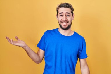 Hispanic man with beard standing over yellow background smiling cheerful presenting and pointing with palm of hand looking at the camera. 