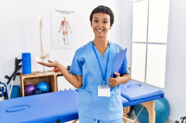 Kısa saçlı, İspanyol bir kadın. Ağrı kesici kliniğinde çalışıyor. Gülümsüyor, elini kaldırıp kameraya bakıyor.. 