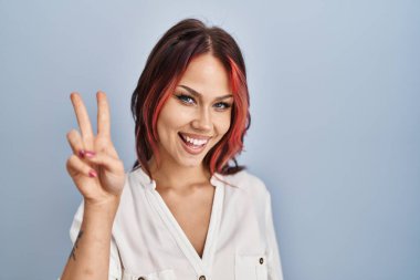 İzole edilmiş arka planda günlük beyaz gömlek giyen genç beyaz kadın kameraya gülümseyen ve parmaklarıyla zafer işareti yapan. İki numara.. 