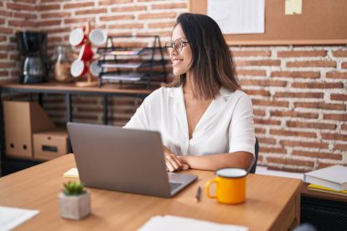 Hispanic young woman working at the office wearing glasses looking to side, relax profile pose with natural face and confident smile. 