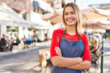 Young hispanic woman waitress standing with arms crossed gesture at coffee shop terrace