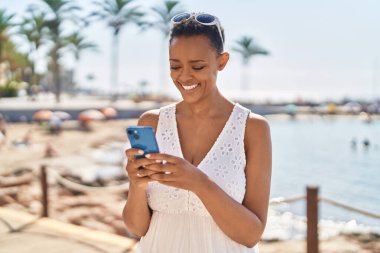 Afrika kökenli Amerikalı bir kadın deniz kenarında akıllı telefon kullanarak gülümsüyor.