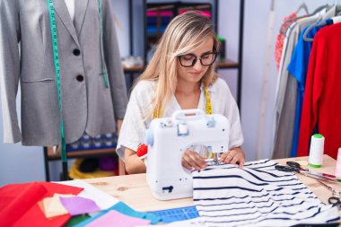 Young blonde woman tailor using sewing machine at tailor shop