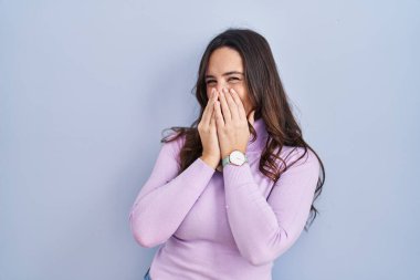 Genç esmer kadın mavi arka planda durup gülüyor ve utanıyor kıkırdıyor elleriyle ağzını kapatıyor, dedikodu ve skandal konsepti yapıyor. 