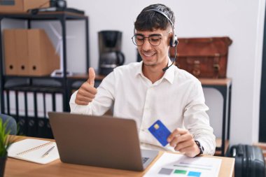 Young hispanic man working using computer laptop holding credit card approving doing positive gesture with hand, thumbs up smiling and happy for success. winner gesture. 