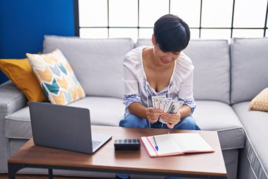 Middle age chinese woman using laptop counting dollars at home
