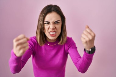 Pembe arka planda öfkeli ve kızgın duran İspanyol kadın öfkeyle bağırırken yumruklarını kaldırıyor ve sinirleniyor. öfke ve agresif konsept. 