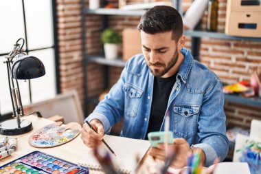 Akıllı telefon kullanan ve resim çizen genç bir İspanyol resim stüdyosunda oturuyor.