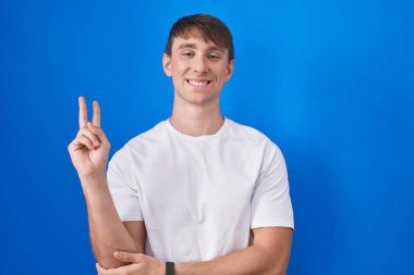 Caucasian blond man standing over blue background smiling with happy face winking at the camera doing victory sign with fingers. number two. 
