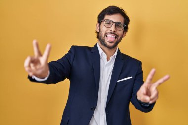 Handsome latin man standing over yellow background smiling with tongue out showing fingers of both hands doing victory sign. number two. 