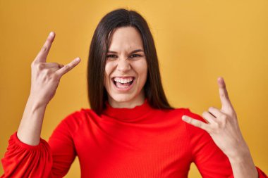 Young hispanic woman standing over yellow background shouting with crazy expression doing rock symbol with hands up. music star. heavy concept. 