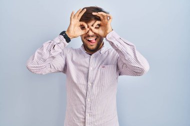 Arab man with beard standing over blue background doing ok gesture like binoculars sticking tongue out, eyes looking through fingers. crazy expression. 