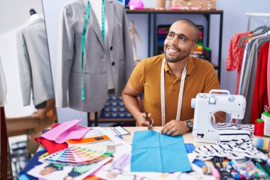 Young latin man tailor smiling confident cutting cloth at atelier