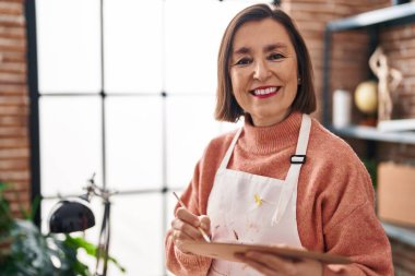 Middle age woman artist holding paintbrush and palette at art studio