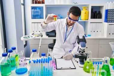 Young hispanic man scientist looking urine test tube writing report at laboratory