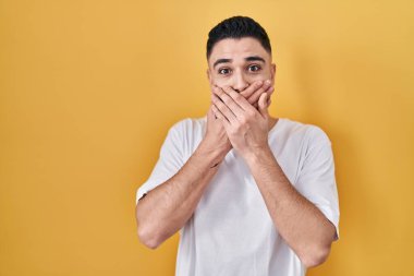 Sarı arka plan üzerine gündelik tişört giyen genç yakışıklı adam yanlışlıkla elleriyle ağzını kapatırken şok oldu. gizli kavram. 