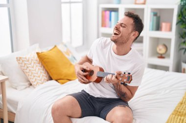 Genç İspanyol adam yatakta ukulele oynuyor.