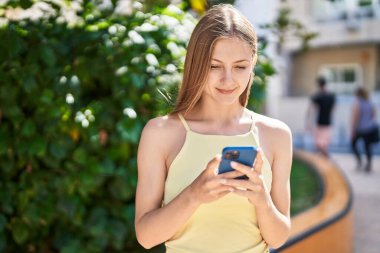 Genç beyaz kadın parkta akıllı telefon kullanarak gülümsüyor.