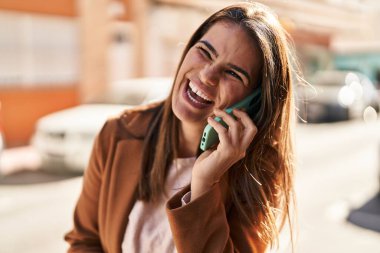 Genç, güzel, İspanyol bir kadın gülümsüyor ve caddede akıllı telefondan konuşuyor.