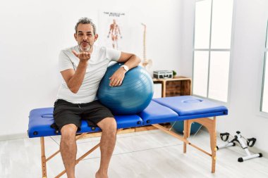 Orta yaşlı İspanyol bir adam ağrı kesici kliniğinde elinde pilates topu tutarken kameraya bakıyor ve havada öpüşürken çok güzel ve seksi oluyor. aşk ifadesi. 