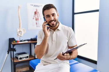 Fizyoterapist üniforması giyen İspanyol genç adam klinikteki akıllı telefondan konuşuyor.